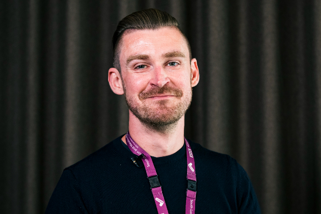 A man is pictured smiling into the camera, standing in front of a dark curtain. He wears a dark top and has a pink strap around his neck holding a work ID. The words "NHS South London and Maudsley" are visible on the strap. He has a stubbly beard and his hair is combed back.