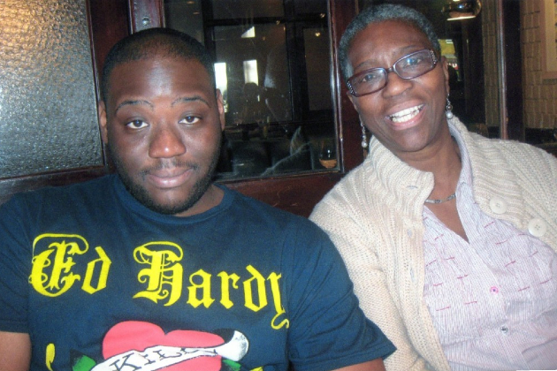 A young man and his mum are pictured together in a pub or restaurant, both smiling into the camera. He wears a dark blue t-shirt with a logo on it and she has a pink top with a cream cardigan over it.