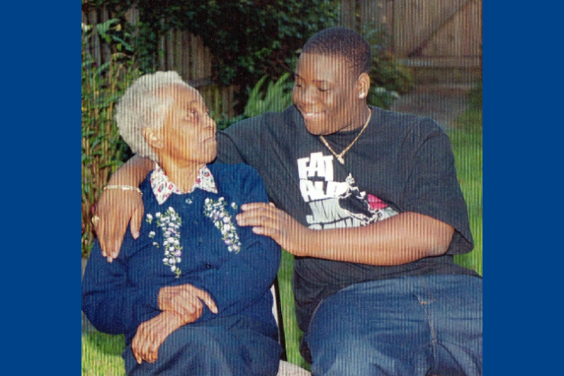 A young man and his grandmother are seated outside. He is on the right and looks down at her with his arm around her and his other hand on her upper arm. She looks up at him. They are both smiling