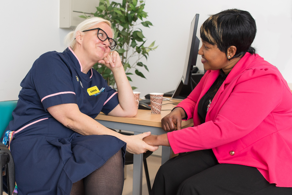 Two nurses sit at a desk facing each other. Anna, on the left, wears her navy nurse's uniform while Nima, on the right wears her own clothes, a bright pink jacket and black clothes. They are good friends, and are holding hands and smiling at each other over cups of tea in paper cups.