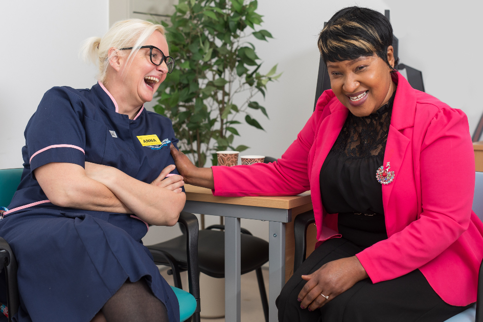 Two nurses sit at a desk facing each other. Anna, on the left, wears her navy nurse's uniform while Nima, on the right wears her own clothes, a bright pink jacket and black clothes. They are good friends, and are laughing together while drinking tea.