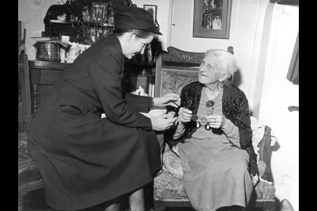 A black and white image from circa 1960s shows a woman district nurse visiting an elderly woman patient in her home
