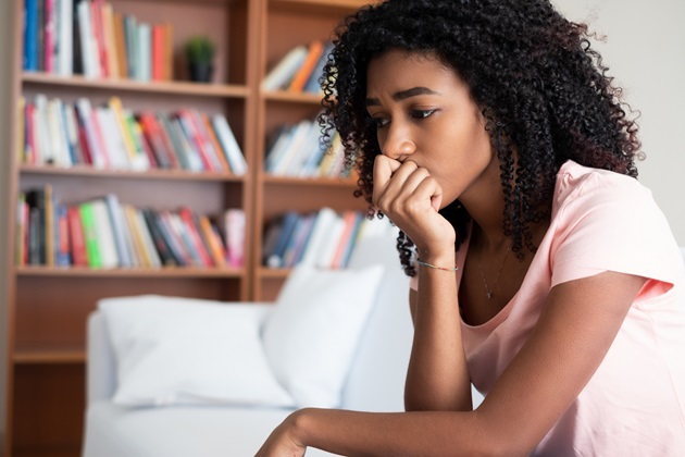 woman sits staring on a sofa resting hand on chin in worry