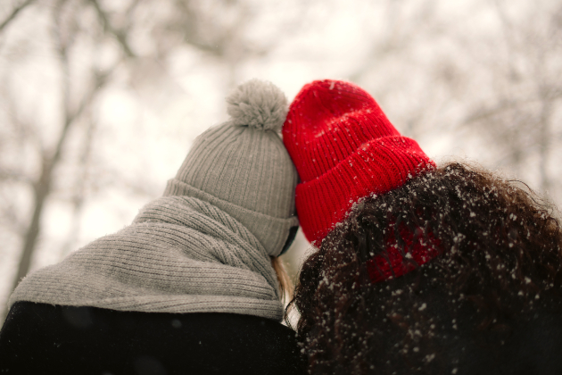 Two people wearing woolly hats are seen from behind. They are touching heads and wrapped up warm with scarves and thick coats. Winter trees with no leaves can be seen in the background