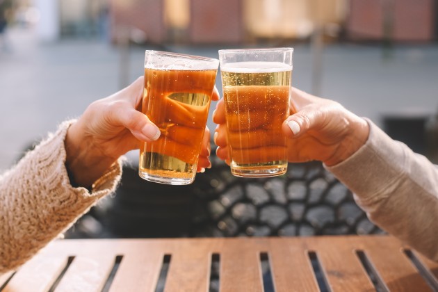 Two glasses of beer are shown being clinked together. You can see people's arms holding the glasses at a wooden table outside.