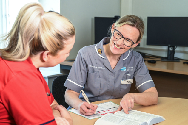 ANP in district nursing Maria Betts sits with a patient in their home. She wears a grey uniform and dark-rimmed glasses with her dark blonder hair tied up. She is smiling at the patient and writing a prescription