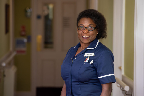 A smiling nurse with glasses, wearing a blue uniform, stands in a care home hallway.
