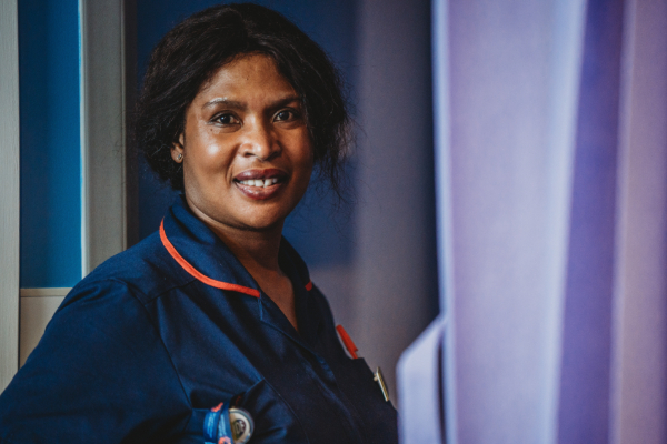 A nurse with a warm smile, wearing a dark blue uniform with a red-trimmed collar, looks directly at the camera in a clinical setting.