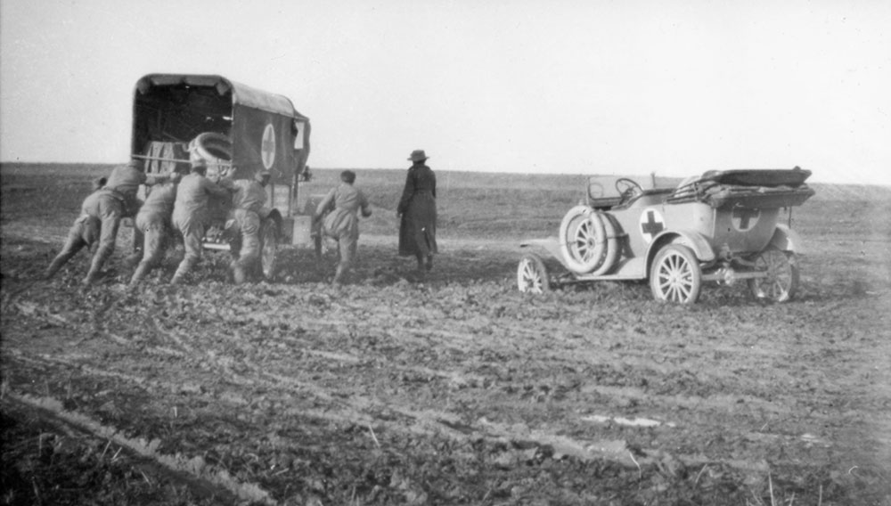 Four soldiers pushing ambulance stuck in the mud, an orderly in cart and an ambulance car in the mud, Romania.