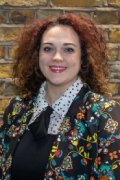 Charlotte Hall, Professional Nursing Committee member for the South West, with long reddish curly hair worn loose, smiling in a very colourful patterned jacket over a black dress with a white collar with black dots on it.