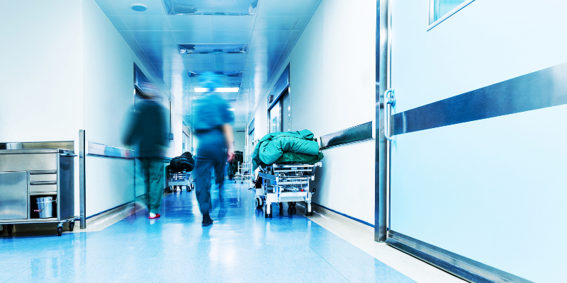 A hospital corridor is shown with medical staff in blurred motion walking down it. Patient beds are seen on either side of the corridor